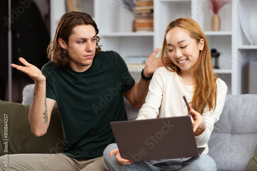 Young Couple Collaborating On Laptop At Home  Man Gesturing With Confused Expression While Woman Smiles  Holding Pen  Positive Work From Home Vibes