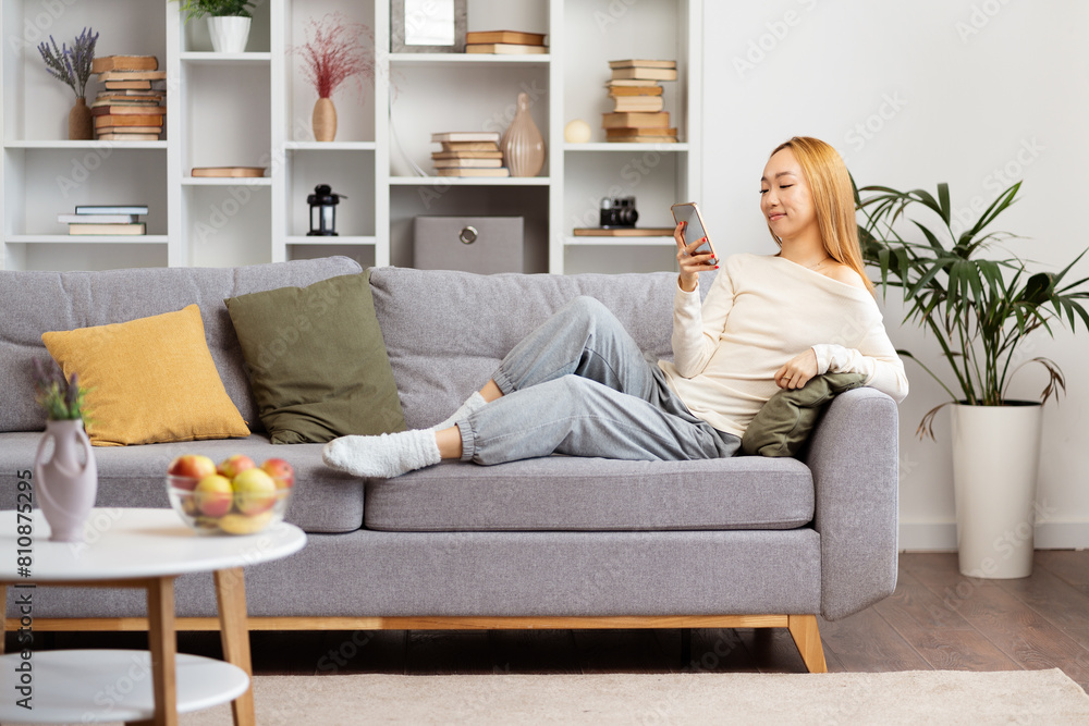 Young Woman Relaxing On Couch At Home With Smartphone, Enjoying Social Media In Cozy Living Room. Millennial Lifestyle, Comfortable Space, Modern Interior Design. Young Woman Relaxing On Couch At Home With Smartphone, Enjoying Social Media In Cozy Living Room. Millennial Lifestyle, Comfortable Space, Modern Interior Design.