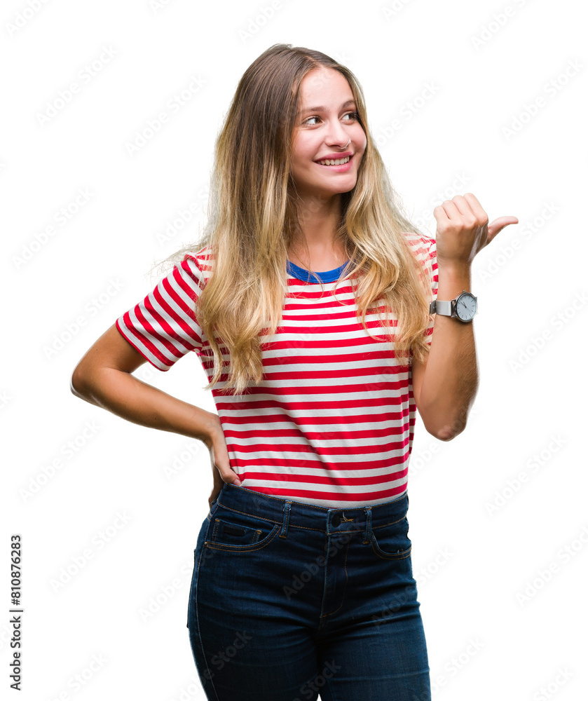 Young beautiful blonde woman over isolated background smiling with happy face looking and pointing to the side with thumb up. Young beautiful blonde woman over isolated background smiling with happy face looking and pointing to the side with thumb up.