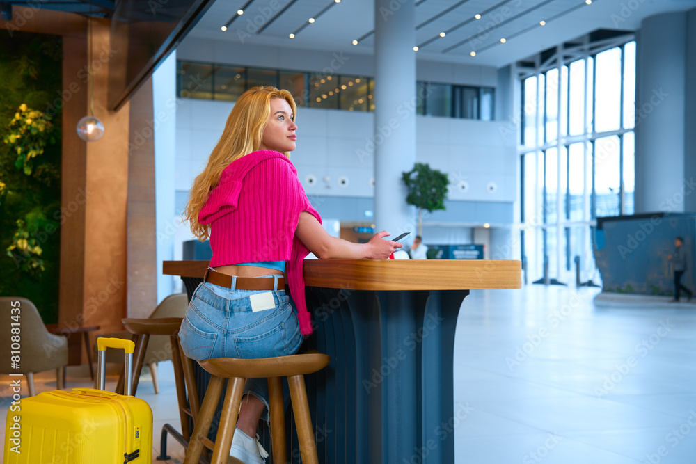 While on vacation, girl is waiting for her flight at airport. She is drinking coffee and using a smartphone, yellow suitcase stands next to her While on vacation, girl is waiting for her flight at airport. She is drinking coffee and using a smartphone, yellow suitcase stands next to her