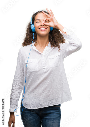 Young hispanic woman wearing headphones with happy face smiling doing ok sign with hand on eye looking through fingers