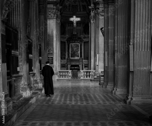 Priest in black robe walking down church nave. Illuminated cross of Jesus.