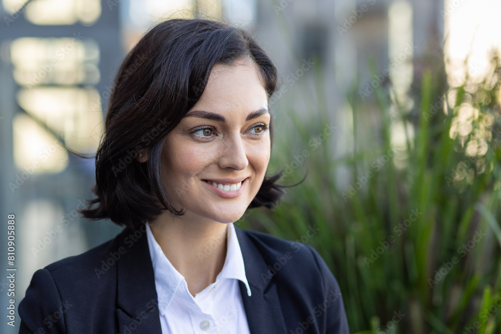 A young smiling business woman is standing outside an office center in a suit and looking away. Close-up photo A young smiling business woman is standing outside an office center in a suit and looking away. Close-up photo