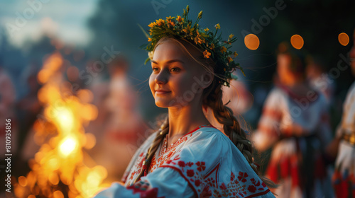 Ukrainian girl in a white embroidered shirt with red floral ornament is dancing around the bonfire, with blonde hair and a green wreath on her head, during the evening at a village festival