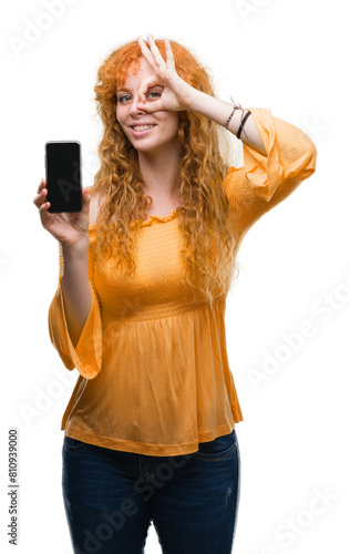 Young redhead woman showing smartphone with happy face smiling doing ok sign with hand on eye looking through fingers