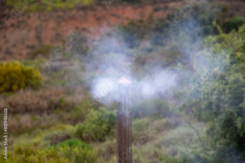Smoke from a wood fire drifts across a field from a metal chimney image in horizontal format