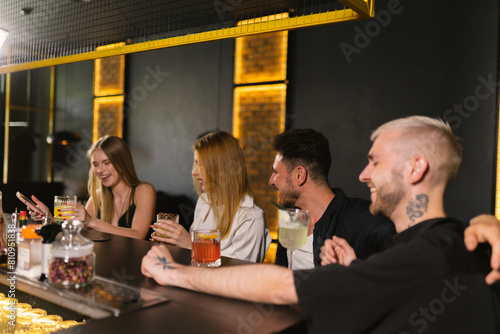 Optimistic friends company at bar drinking alcoholic beverages and laughing out loud. Blonde ladies with evening makeup sit near solid men with tattoos in black shirts