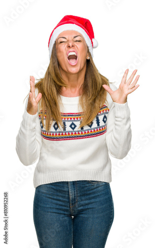 Middle age adult woman wearing winter sweater and chrismat hat over isolated background crazy and mad shouting and yelling with aggressive expression and arms raised. Frustration concept. photo