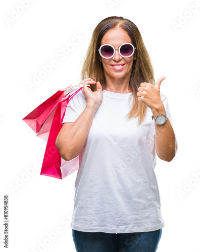 Middle age hispanic woman holding shopping bags on sales over isolated background pointing and showing with thumb up to the side with happy face smiling