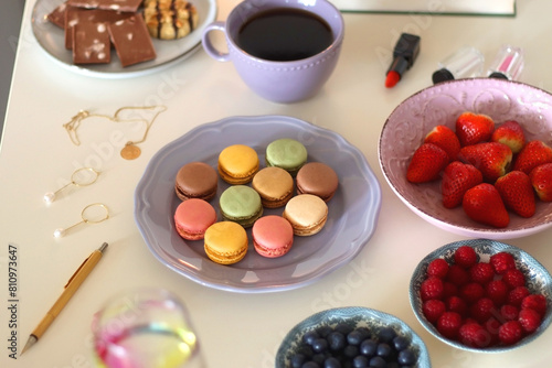 Plate of pastel macarons, cookies and chocolate, cup of tea of coffee, glass of bubble water, various berries, books and accessories on the table. Selective focus, pastel colors.