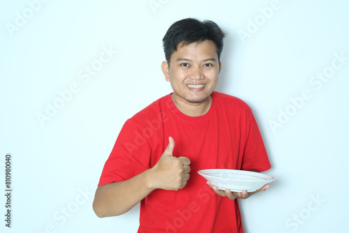 Young Asian man give thumb up and showing enthusiastic expression while holding empty dinner plate. Wearing red a shirt photo