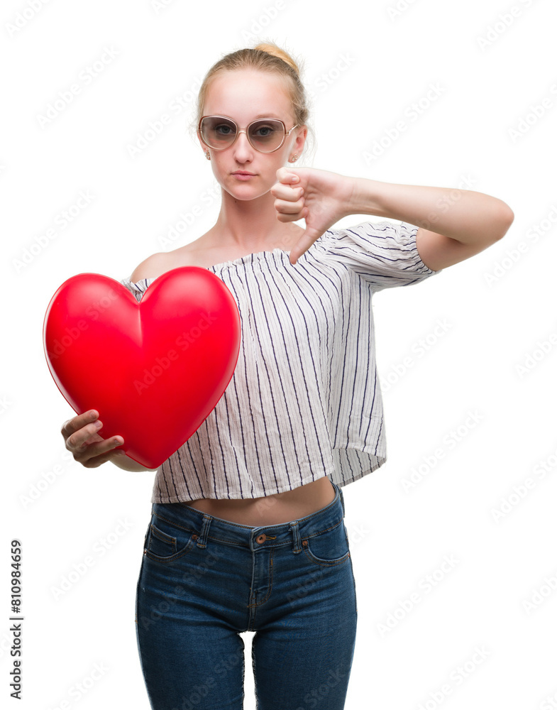 Blonde teenager woman holding red heart with angry face, negative sign showing dislike with thumbs down, rejection concept Blonde teenager woman holding red heart with angry face, negative sign showing dislike with thumbs down, rejection concept