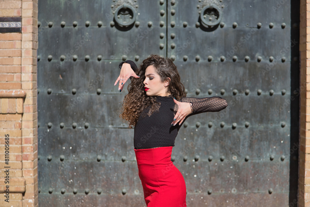 Fototapeta premium Portrait of young, beautiful, brunette woman in black shirt and red skirt, dancing flamenco with old, black metal door in the background. Flamenco concept, dance, art, typical Spanish.