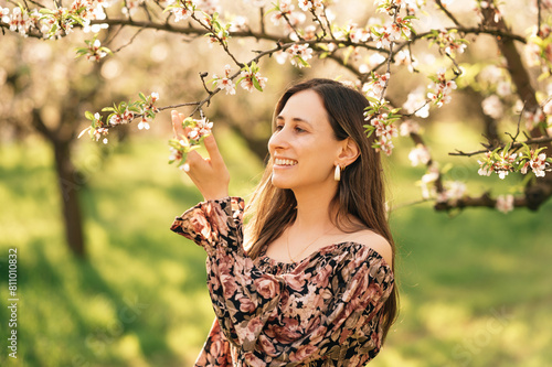 Outdoors close up portrait of a smiling woman touching almond tree in blossom.