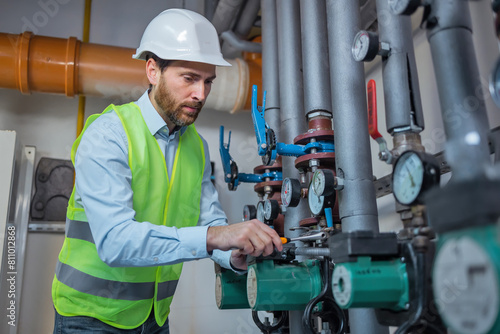 Plumber in protective helmet tightening nut in the boiler room. Repairman fixing a hot water pipe in the technical area of heating system. Adjusting water flow through pipes. repair service. © Ivan