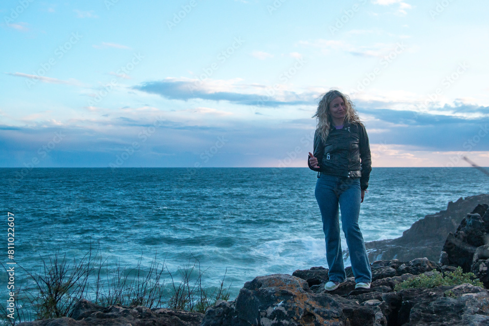Adult blonde woman dressed in jeans and leather jacket photographed during sunset with the ocean behind her Adult blonde woman dressed in jeans and leather jacket photographed during sunset with the ocean behind her