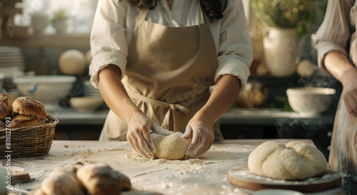 A woman in an apron kneading dough on the table, with her hands covered by flour and she is looking at it intently, while behind him there's another person making bread in the background