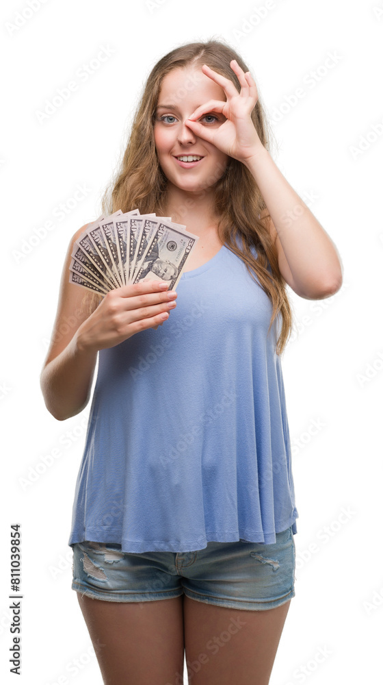 Young blonde woman holding dollars with happy face smiling doing ok sign with hand on eye looking through fingers Young blonde woman holding dollars with happy face smiling doing ok sign with hand on eye looking through fingers