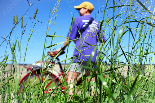 A girl in a cap and a blue T-shirt holds a bicycle, a view through and through the tall grass under a clear blue sky.