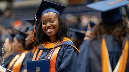 Close up african american college or university graduation students in black and orange gown and graduate cap smiling at camera at the foreground holding her blue diploma