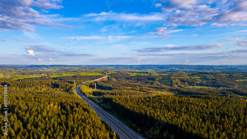 This aerial photograph captures the sweeping view of the E42 highway as it cuts through the verdant landscape of the Hautes Fagnes area near Emmels. The lush green forests dominate the scene  with