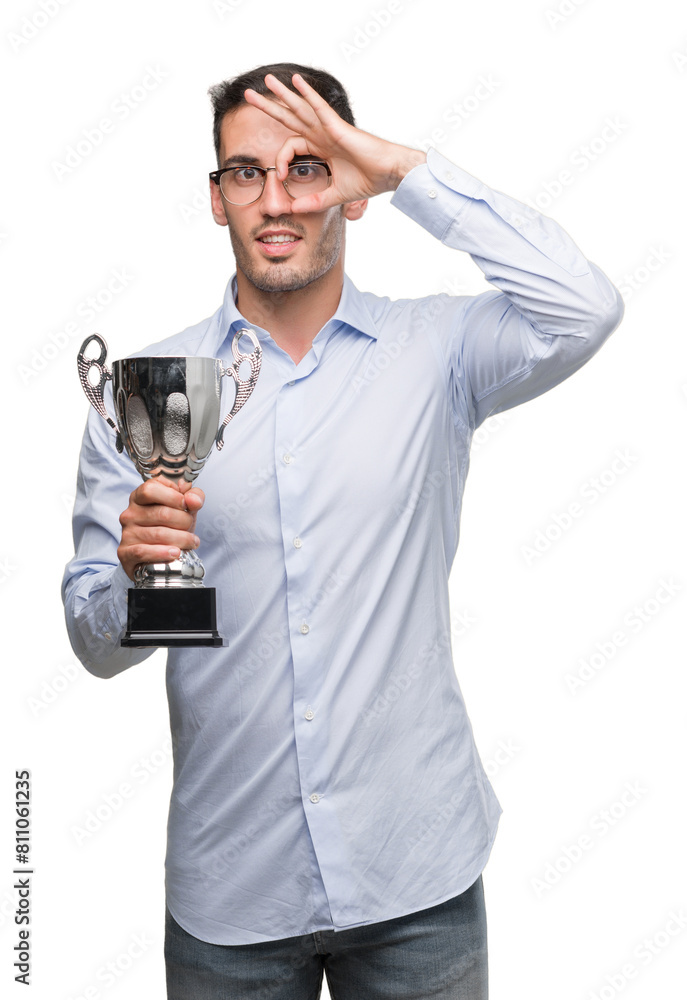 Handsome young man holding trophy with happy face smiling doing ok sign with hand on eye looking through fingers Handsome young man holding trophy with happy face smiling doing ok sign with hand on eye looking through fingers