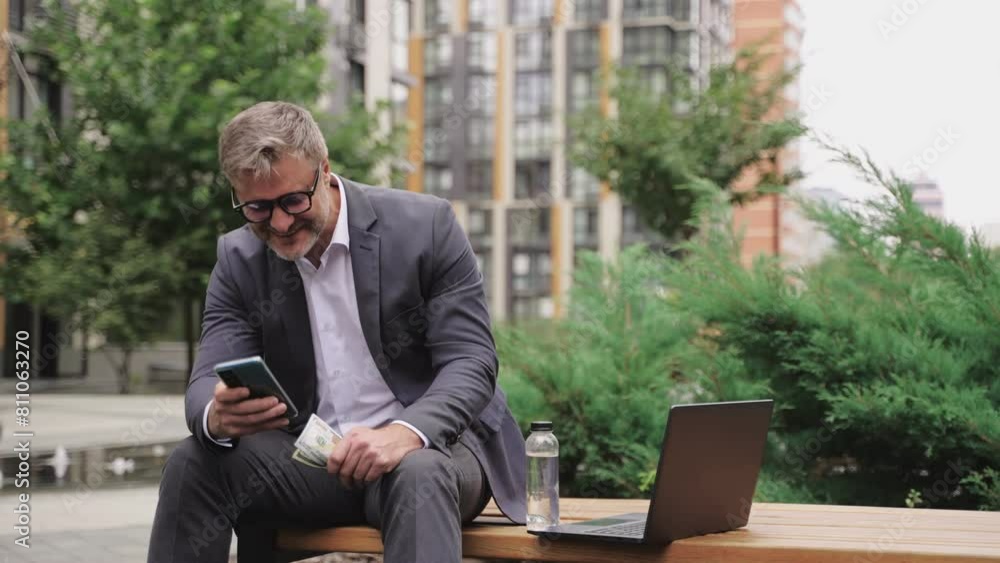 Happy adult man using smartphone and money, sitting on the bench. Real time concept Happy adult man using smartphone and money, sitting on the bench. Real time concept