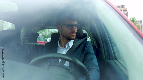 Serious young confident man in glasses driving car in the city while looking around. Transport, business and people concept. Real time