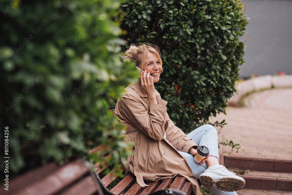 Fototapeta premium Young attractive blonde woman talking smartphone during coffee break sitting on bench in city park
