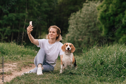 A woman takes a selfie with her beagle dog while relaxing in the park on a summer day. Social media concept, joyful moments with pet, friendship