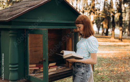Red-haired teenage girl chooses a book from the free open public library at city park