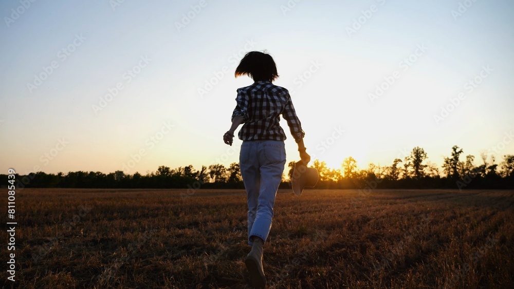 Fototapeta premium Female farmer running through the barley plantation at sunset. Agronomist jogging among wheat meadow at dusk. Beautiful scenic landscape. Concept of agricultural business. Close up Slow motion