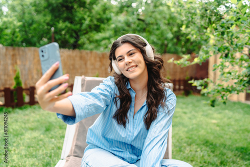 One young caucasian woman is taking selfies while relaxing in easy chair in her backyard and listening to music on wireless headphones