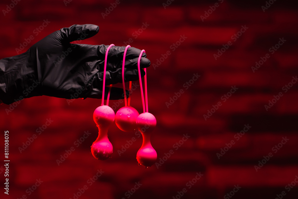 Obraz premium Woman's hand in black latex gloves holds pink Kegel balls and Ben Wa balls on a red background. Vaginal balls in a woman's hand. Sex toys Geisha Balls, Products for intimate stores