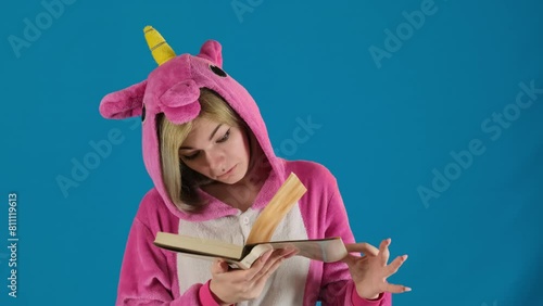 Female teenager in unicorn pajamas opens book turning page on blue background. Girl in hood expresses unwillingness to read hometask photo
