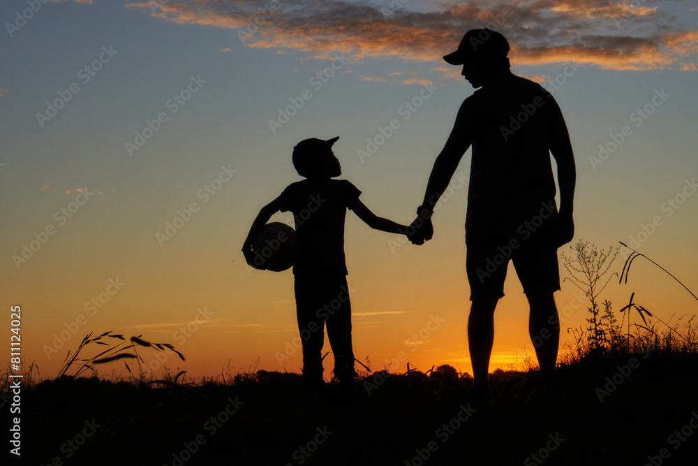 silhouette of father and son hand in hand with a ball at sunset in the field