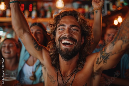 Tattooed man smiling widely, celebrating with raised hands among friends in the lively ambiance of a bar or party