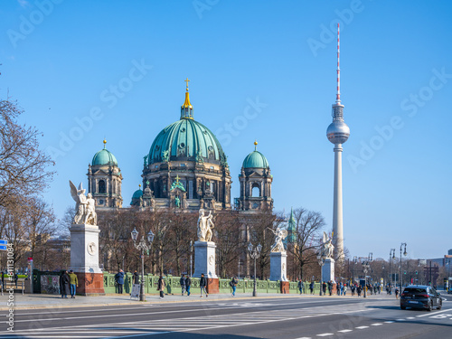 The Berlin Cathedral, or Evangelical Supreme Parish and Collegiate Church, basking under a clear blue sky with Berlins iconic TV Tower in the background and pedestrians nearby. Berlin, Germany photo