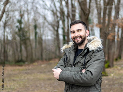 Young and handsome bearded man looking at camera and smiling in park, woodland. A man wearing a coat in cold weather