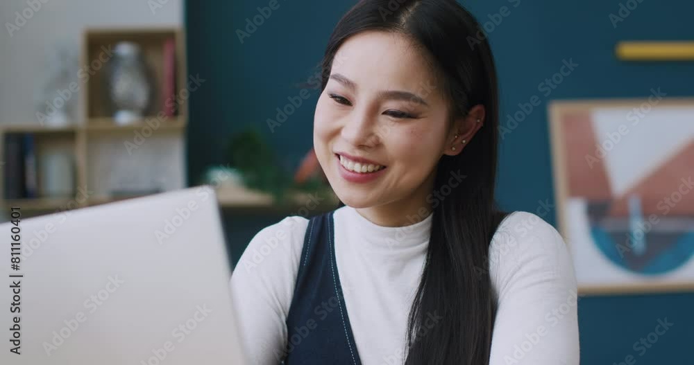 Happy Chinese woman using laptop while spending time at home. Looking with focus at display. Smiling with joy after getting new message on her mail. Enjoying communication with friends online.