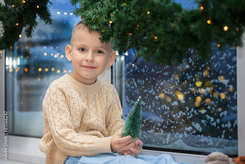 Little happy boy sits on the windowsill of a snow-covered window with Christmas tree decoration. Child in the Christmas room.