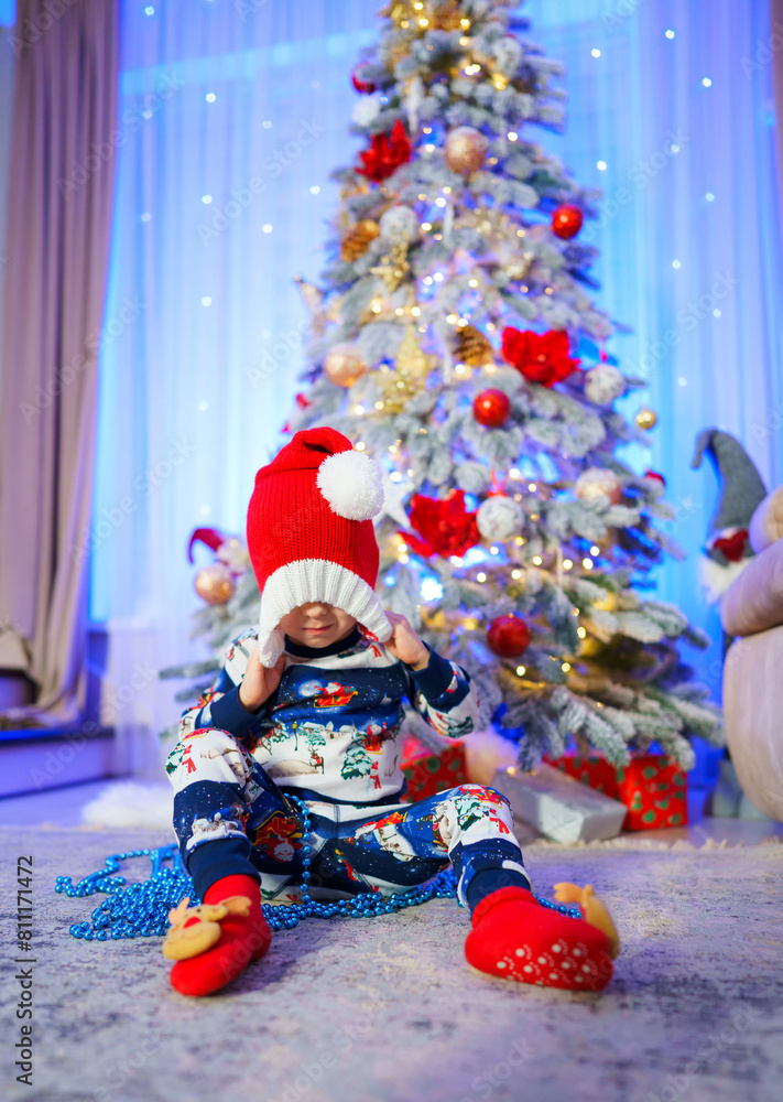Fototapeta premium A young child is sitting in front of a Christmas tree, wearing a red hat and a blue and white snowsuit