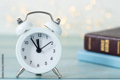 Alarm clock with holy bible and notebook on wooden table with white bokeh light background. Close-up. Christian biblical concept of time, patience, Scripture study, and prayer.