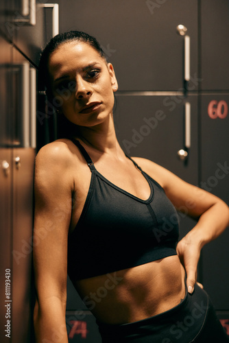 Portrait of a muscular fit girl, leaning on the locker at the gym, posing for the camera, dressed in a sports bra. (ID: 811193430)