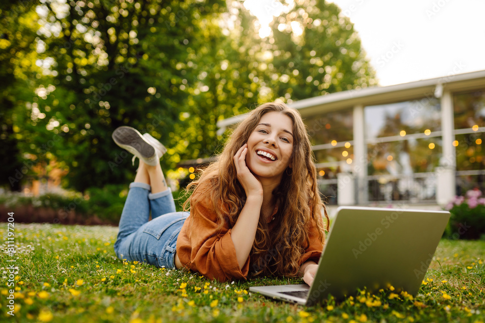Portrait of attractive young woman sitting on green grass in park while using laptop and wireless earphone. Freelancer, education, online, technology concept.