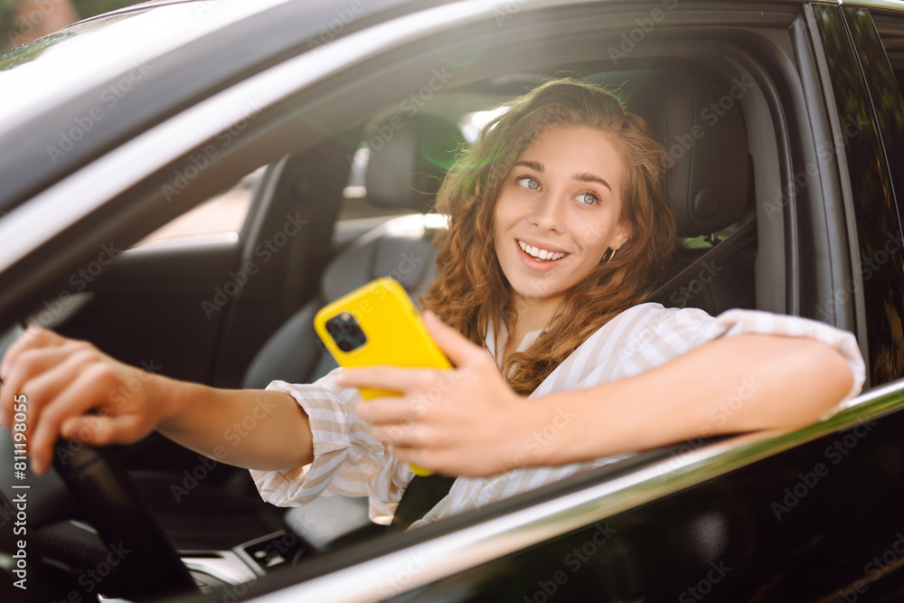 Young woman sitting in a car in the driver's seat looking into a smartphone, paying for parking and navigating in the city. Leisure, travel, technology.