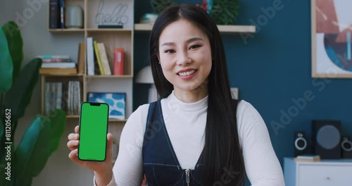 Positive Asian girl holding phone with green screen in her hands. Cute smiling and looking at camera. Holding up big finger while recommending smartphone with chroma key for sale or use. photo
