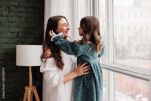 Mother's day. Happy young mom embraces with her little adorable daughter by window at home. Trust, support and love between mommy and child. Loving family enjoy tender sweet moments. Children day.