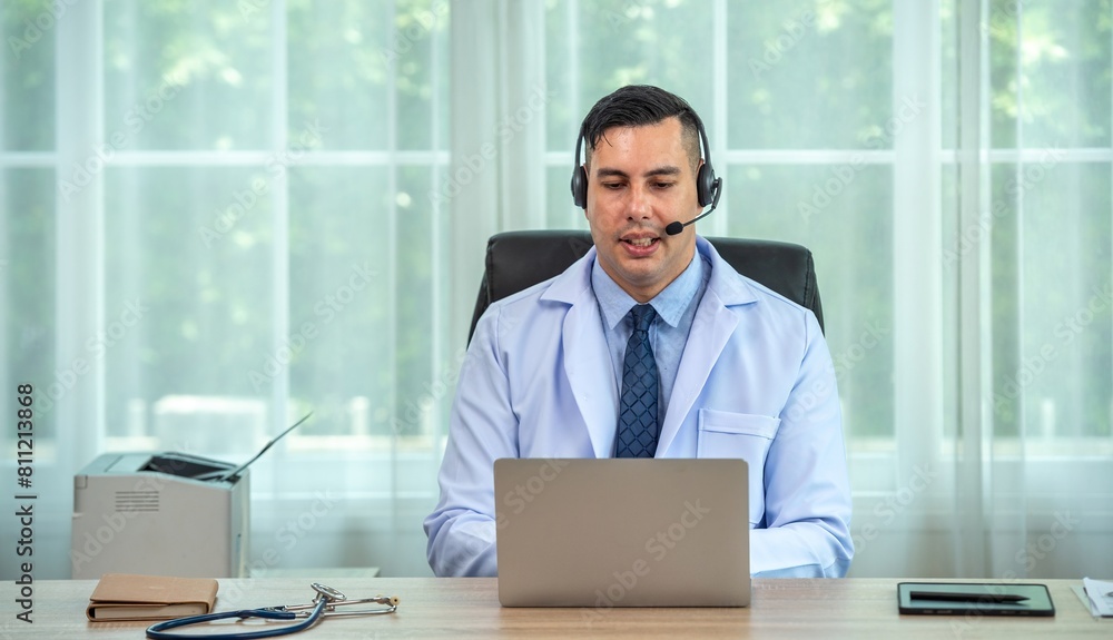 A doctor wearing a headset is sitting at his desk and looking at his laptop. A doctor wearing a headset is sitting at his desk and looking at his laptop.