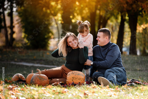 Thanksgiving day. Happy mom, dad and little child daughter having fun sit on grass with pumpkins at autumn park, enjoying spend time together at weekend. Parental care and happy carefree childhood